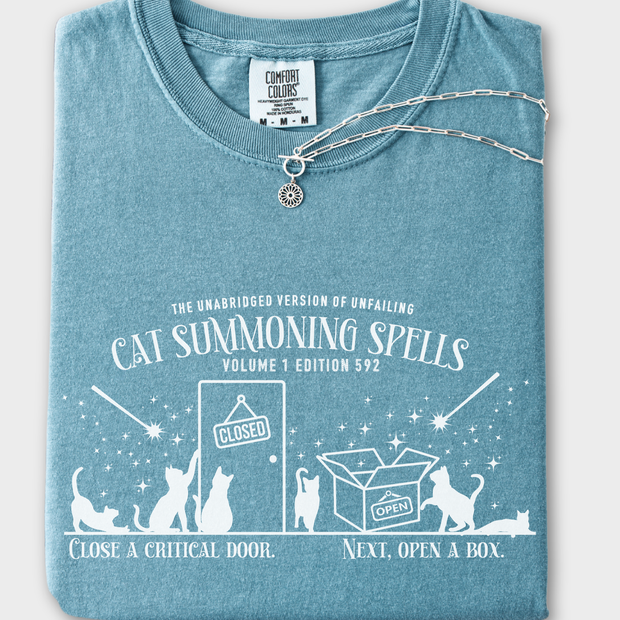 Minimalist cat-themed T-shirt featuring a 'Cat Summoning Spells' book graphic, where wands magically summon cats to doors and boxes.